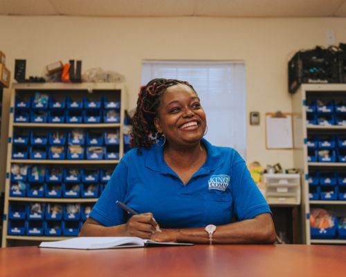 A smiling woman in a polo shirt sitting at a table and writing in a book.
