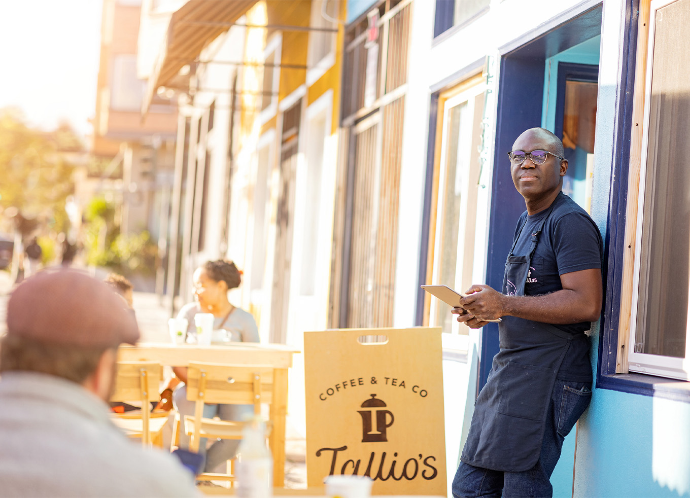 A man leaning against the outside wall of a coffee shop, looking off into the distance. Tallio's sign next to him.